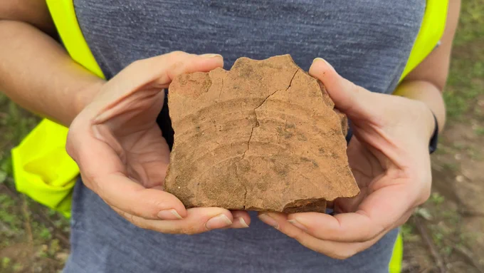Volunteer holding an piece of thick orange tile with three curved fingermarks on one side - probably the base. Fragment is slightly larger than hand-sized.