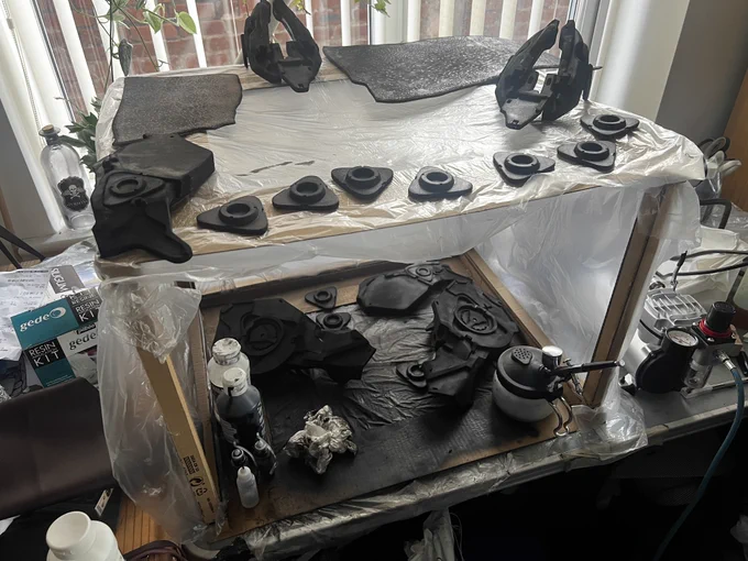 A collection of armour made of foam and primed sitting in carboard and plastic sheet cube. There is an airbrush in alongside and the base is covered in paint