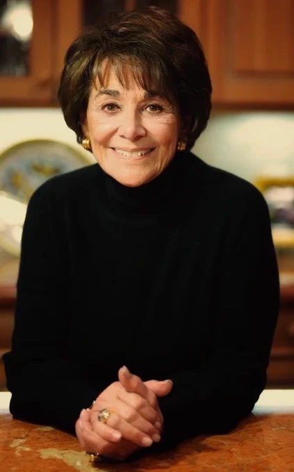 Congresswoman Anna Eshoo wearing a black sweater, gold hoop earrings and a gold ring sits with her hands folded on top of a granite brown kitchen counter. The maple wooden cabinets are in the background.