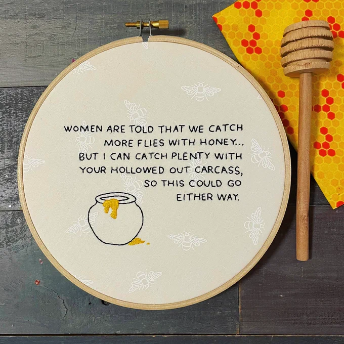 An embroidery hoop with white fabric, covered in subtle white bee print. Embroidered on it in black are the words “women are told that we catch more flies with honey…but I can catch plenty with your hollowed out carcass so this could go either way” beside a stylised image of a pot of honey