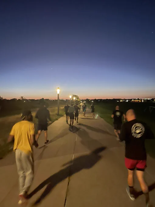 Group of men in pre-dawn jogging away from view down a light lined path in a park.