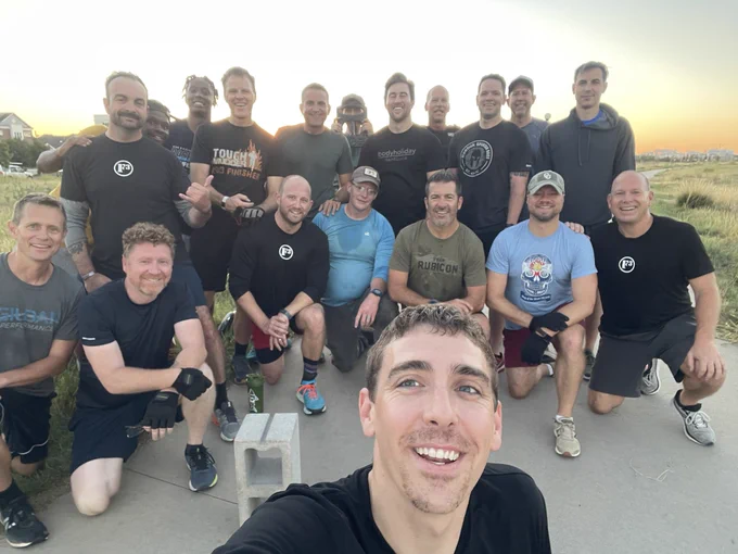 19 men in a group smiling at camera with sunrise in the background.