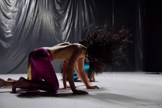 Three topless women in bright trousers crawl on a stage, their dark hair flicks upwards