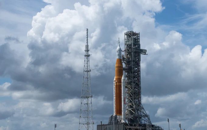 The Space Launch System rocket stands upright on the launchpad. The background is the sky dominated by puffy cumulus clouds. The rocket has an orange central fuel tank with two white rocket boosters on either side. The boosters have the word NASA painted vertically on them in red in the iconic "NASA Worm" typeface. The typeface resembles a worm. Photo credit: NASA/Joel Kowsky