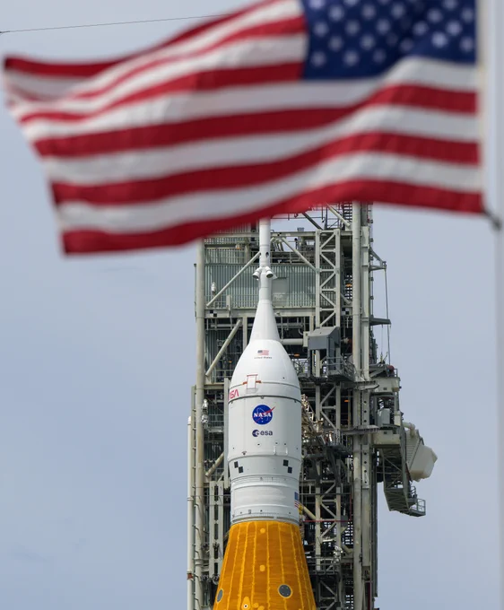 The Space Launch System rocket and Orion Spacecraft are seen with the American flag in the foreground. The sky is grey in the background. 