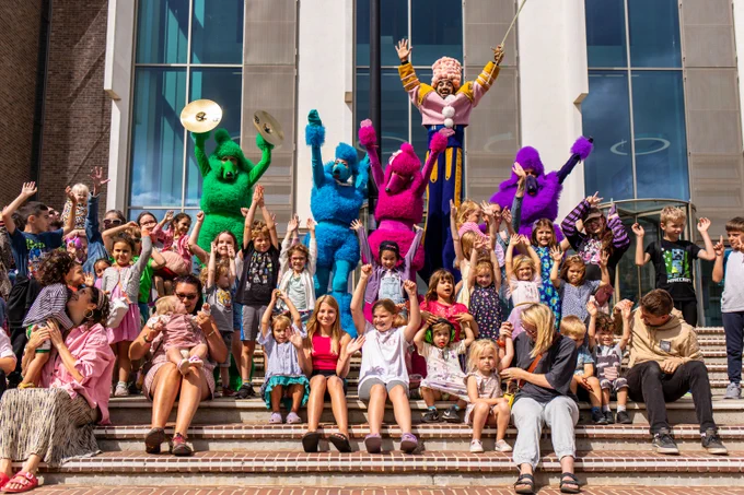 Children, their families and the cast of the Royal Poodle Parade team wave their hands in the air for a photo outside the University of Kent Templeman Library 