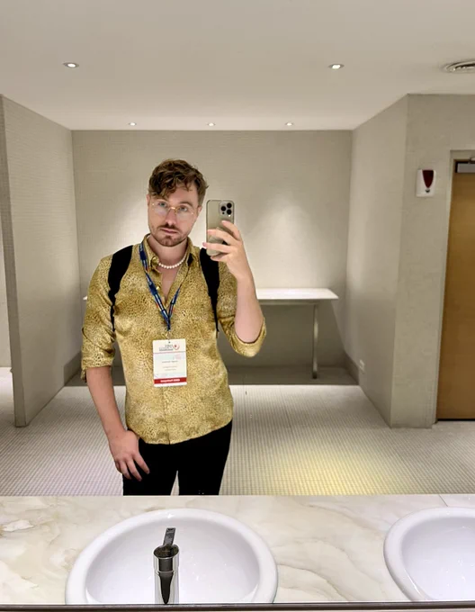 A picture of me on a bath room mirror. Sinks are in the foreground. I am wearing a gold and black cheetah print shirt with a necklace of pearls. I have stubble on my face and I am wearing glasses, my hair is slightly curly and a brown blonde. Around my neck as a lanyard for the conference and on that is a badge that says they/them