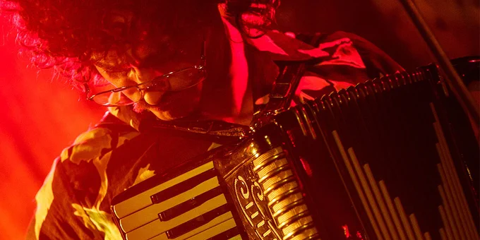 A close-up of Daniel Radcliffe as Weird Al Yankovic in Weird: The Al Yankovic Story. He is looking down while playing the accordion passionately under red stage lights.