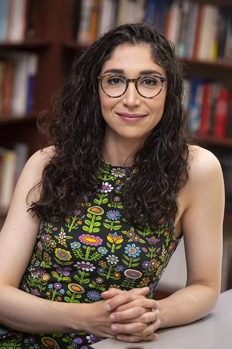 Francesca Zaffora Blando is pictured with her hands folded in front of her, leaning against a table. She is wearing glasses and a sleeveless black dress with multicolored flowers on it.