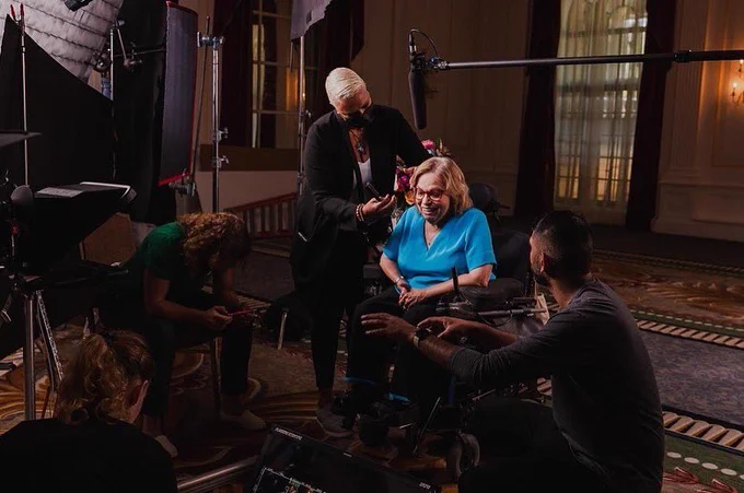 An image of Judy on set getting her hair touched up by a makeup artist. Around Judy are three other people, lights, screens, and a boom microphone.