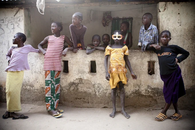 Colour photography print of group of young kids, hanging out in their compound, one girl wearing a mask, looks at the camera.  Image © Jason Florio, West Africa.