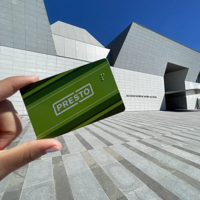 A hand holding out a PRESTO card in front of the Aga Khan Museum