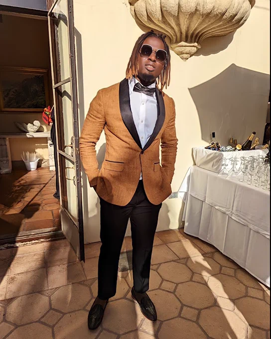 Kevin posing with his hands in his pocket, wearing a orange and black suit, bowtie, and sunglasses.