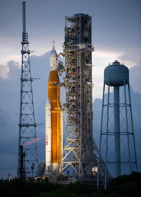 The Space Launch System rocket and Orion spacecraft sit at Kennedy Space Center with dramatic clouds behind in shades of light blue and grey.