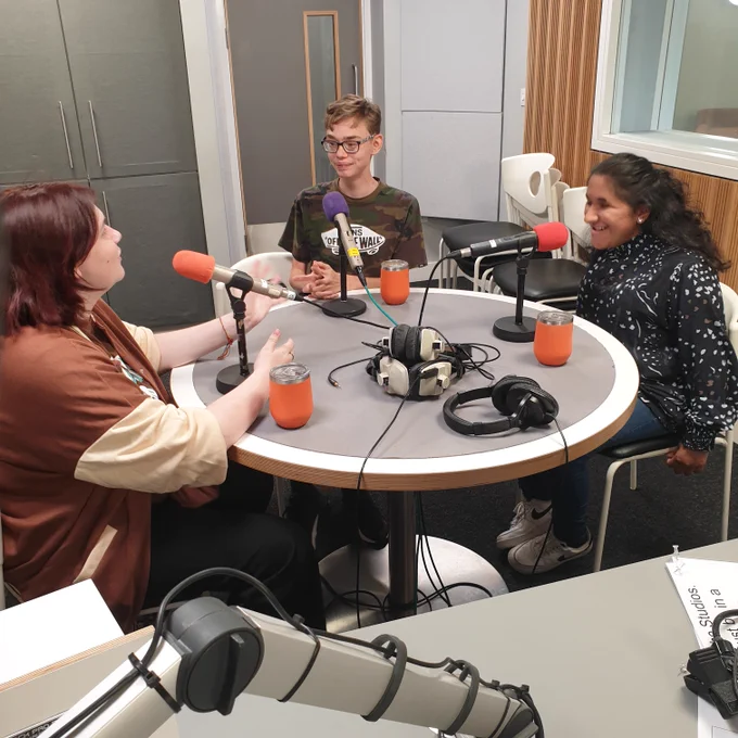 Three people sat around a table chatting and laughing in a radio studio
