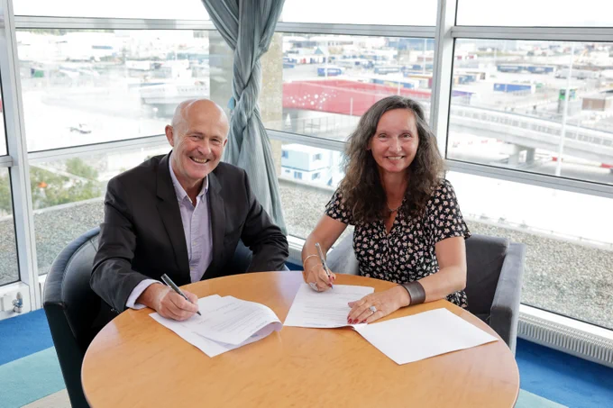 Two people sit at a round table with documents in front of them. They both hold pens as though they are poised to sign the documents. Behind them are windows through which part of Dublin Port is visible.