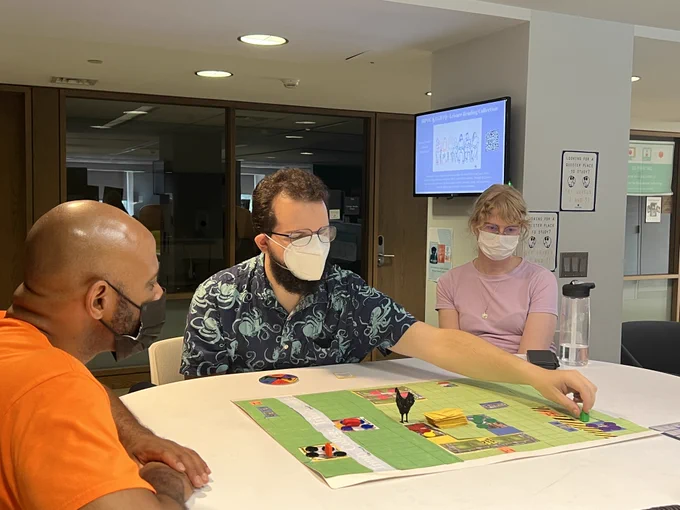 Thee people sitting around a game board that is predominately green. The person in the center is reaching forward to move their game piece.