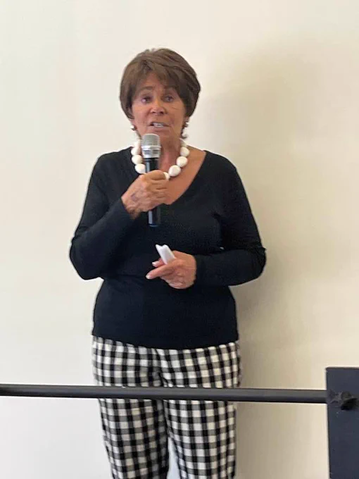 Congresswoman Anna Eshoo is speaking holding a microphone. She's wearing a chunky necklace, black sweater and black and white checked pants.