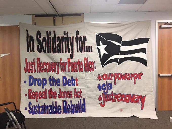 Banner that reads "Solidarity for... Just Recovery for Puerto Rico: - Drop the Debt, Repeal the Jones Act, Sustainable Rebuild, #OurPowerPR #CJA #JustRecovery" together with the black and white puerto rico flag