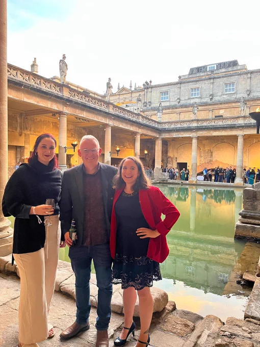Two women and one man stand in front of a large, ancient Roman bath filled with hotspring water and lined with sandstone columns. They are dressed semi formally and smiling while looking at the camera