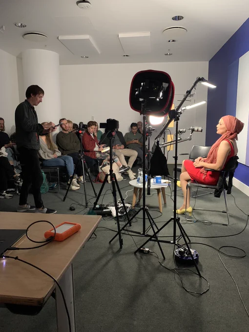Sadia and Leon’s talk on video for podcasts. Leon is setting up cameras and lighting as Sadia sits at a table with a mic near her. We can see a crowd and a purple and white wall