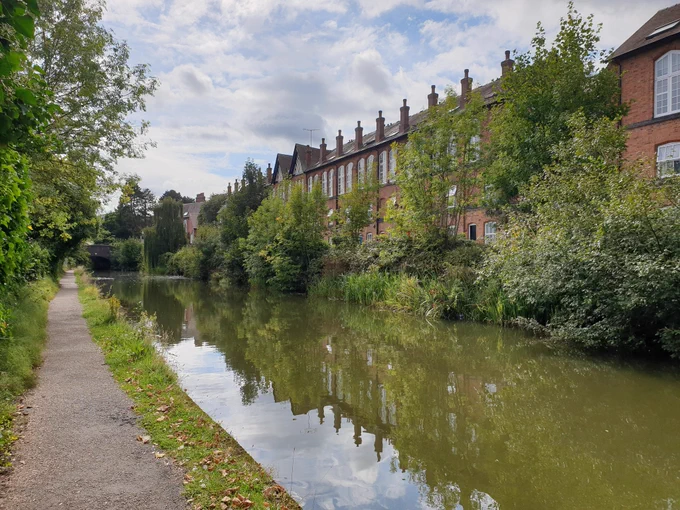 Canal with well surfaced tree lined towpath. Other side lined by older, once industrial buildings now converted to residential. Buildings reflected on water surface.