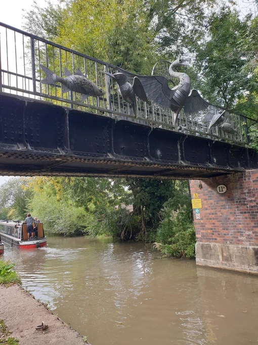 Canal with narrow boat heading away from shot.  Metal Bridge over canal with metal sculptures of birds and fish.