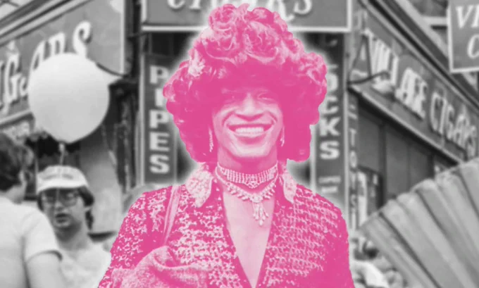 Marsha P. Johnson stands in front of a corner store, a person holding a balloon behind her to the left. Most of the photo is in black and white, but Marsha is bubblegum pink.