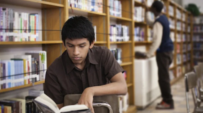 Candid photo of a young teenage male student with medium brown skin and short black hair sitting backwards on a chair in a school library. Shelves of book fill the background and there is another person, perhaps a librarian, with black skin and black hair reaching into the shelves.