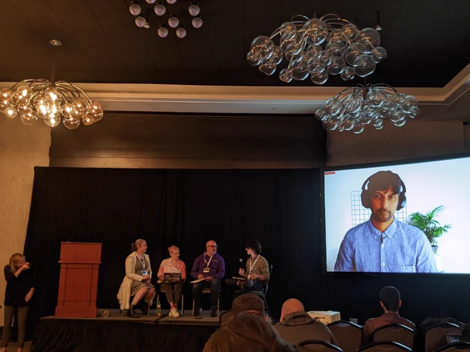 Photo is from the audience point of view of a panel of people. From the left is a white-presenting woman in black doing interpretation, then Silona a white woman in a white & black ensemble with a 3D printed octopus necklace. To the right of Silona is Eriol, who is a white non-binary person in pale pinks and white with dark grey, followed by Sean, a bald white man with black glasses in dark purple and blue, and last on stage is Django, a white-presenting person whose pronouns I missed, dressed in a button up shirt with a dark pattern on a lighter tan. Further to the right on the big screen is Karthik, a Brown man in a blue and white thin pinstripe shirtsleeve button down shirt, wearing black headphones against a neutral virtual background. The room around shows the panel is in a modern hotel ballroom.