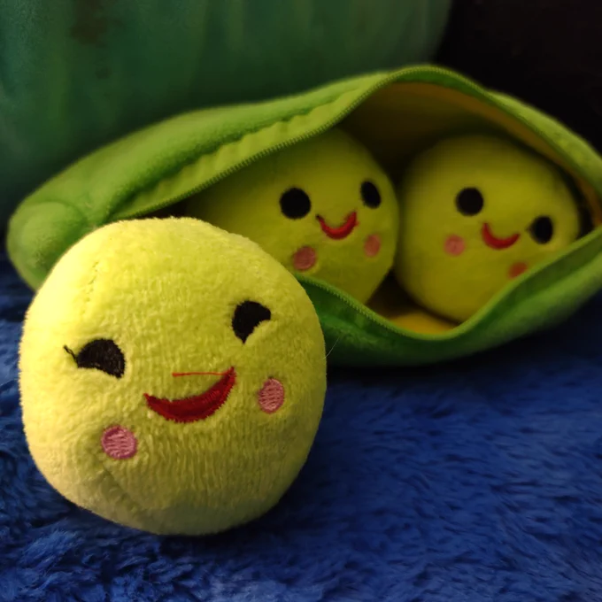 A green cloth pouch that looks like a pea pod, with 2 smiling green peas sitting inside, and 1 smiling green pea sitting out side of the peapod.