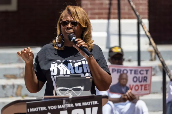 LaTosha Brown stands at the bottom of a set of concrete steps at a podium, speaking into a handheld microphone. Someone sits behind her on the steps holding a sign with a photo of former U.S. Rep John Lewis and the words "Protect our vote!"