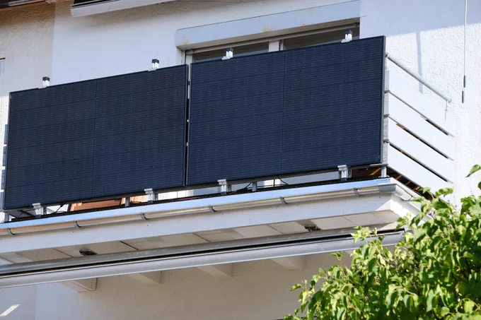 Zwei dunkle Solarmodule hängen an einem Balkon. Die Sonne scheint darauf, im Vordergrund ein grüner Braum. 