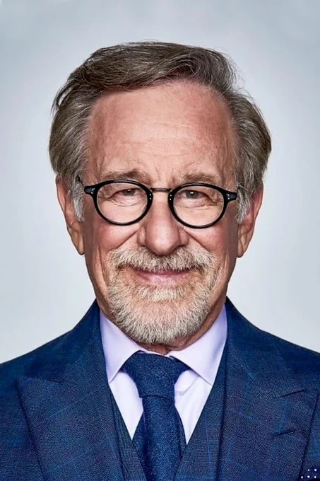 A portrait of Steven Spielberg wearing a crisp blue suit and circular black glasses. He smiles while looking directly into the camera in front of a white background.