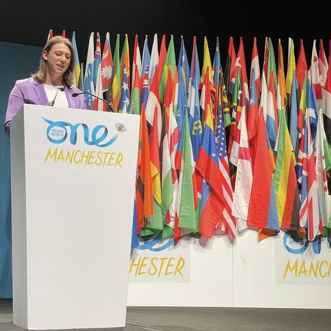 Vermont State Representative Taylor Small is delivering remarks from behind a white podium with the One Young World Manchester logo on the front and a collection of the world’s flags behind her. She is wearing a purple suit and has medium length brown hair.