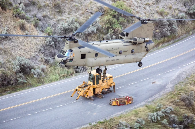 a large helicopter hovering above a large construction vehicle on a road. A person in military uniform is beneath the helicopter working to strap the construction vehicle to the helicopter for transport.