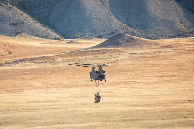 a large helicopter carrying a large construction vehicle flying through the air. A grassy meadow and mountains are seen in the distance.