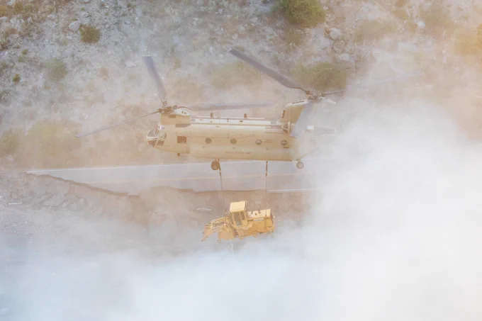 a large helicopter carrying a large construction vehicle flying through a canyon. The propellers are kicking up a bunch of dirt and dust into the air.