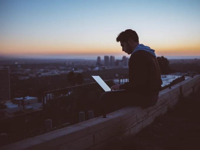 Photo showing man sitting on a concrete brick with opened laptop on his lap captured by Avi Richards and taken from unsplash.com 