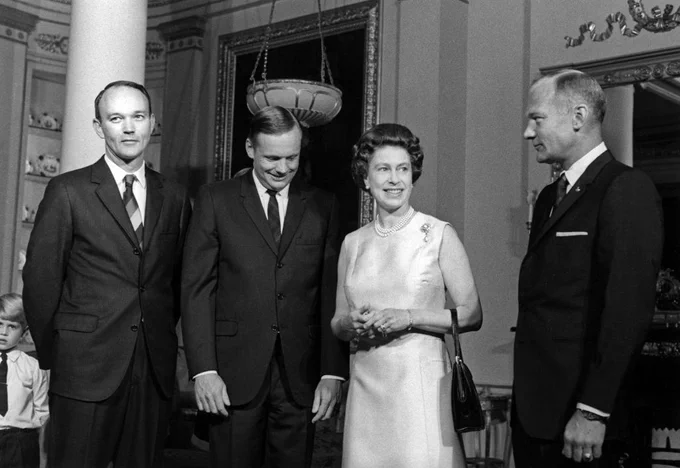 This black-and-white image depicts a 1969 event at Buckingham Palace where (left to right) Michael Collins, Neil Armstrong, Queen Elizabeth II, and Buzz Aldrin met as part of the Apollo 11 Goodwill Tour. The NASA astronauts wear dark suits and slim ties. The queen appears in a light-colored sleeveless dress, pearls, and a brooch.