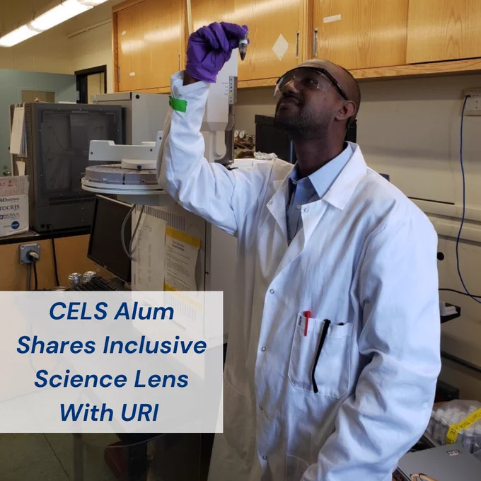 Image of Asta Habtemichael wearing safety glasses, purple latex gloves, and a white lab coat, holding something above eye level and looking at it. Wooden cabinets and tan walls in the background of the image. A small white box in the bottom left corner with navy writing reading “CELS Alum Shares Inclusive Science Lens With URI”