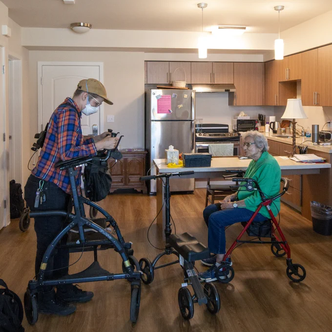 Photo of a consumer and CIL's Assistive Technology Coordinator in an apartment with two different rollators and a knee scooter. The consumer is sitting on a red rollator and CIL's Assistive Technology Coordinator is testing a black rollator.