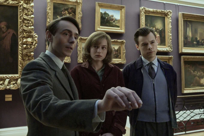 David Dawson, Emma Corrin and Harry Styles stand studiously in front of a wall with framed artworks in the film My Policeman.