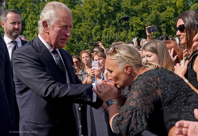 A woman, surrounded by a crowd of people, kisses the hand of Britain's King Charles III.