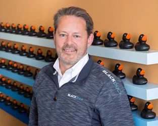 Portrait of Lou Shipley '85. Background shows shelves with five rows of black rubber ducks.