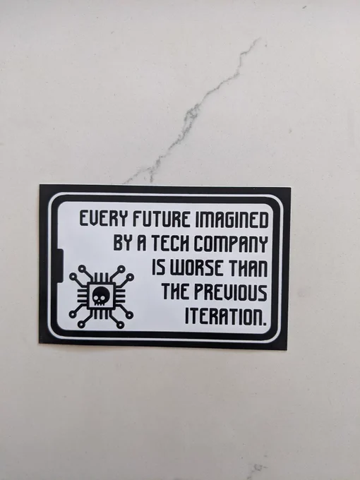 A sticker which reads, in computer-like font, "Every future imagined by a tech company is worse than the previous iteration." It is accompanied by a stylized illustration of a computer chip with a skull in the middle. The sticker is atop a marble countertop.