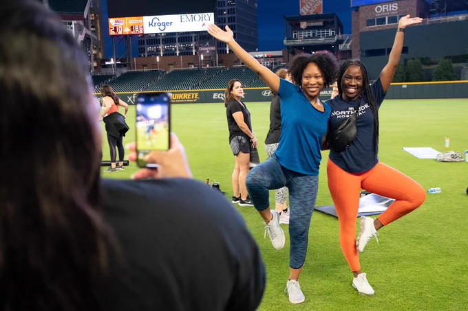 Two guests pose while another takes their photo with a cell phone after yoga class on field at Truist Park