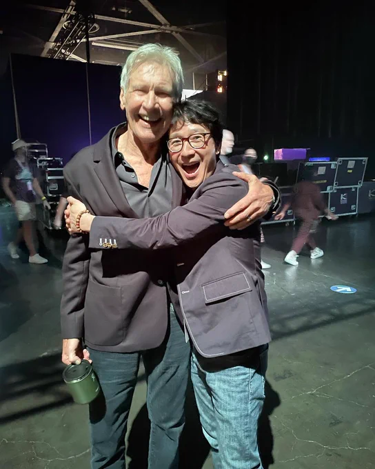 A very cute photo of actor Ke Huy Quan hugging Harrison Ford in absolute joy. Ke's arms are wrapped around Harrison. They are in back stage setting and wearing dark greyish jacket and jeans. Harrison is holding some kind of green tin.