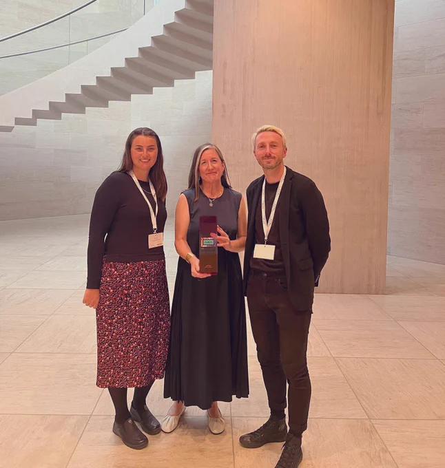 Image Description: Three people stand smiling together in the foreground and centre of the photo. The floor and background of the photo is neutral and white and a staircase can be seen. From Left to Right is - Orla Dalby wearing a black top and long red skirt; Fiona Kearney wearing a dark long dress holding the Art Museum Award; Tadhg Crowley wearing a black top and black jeans. 