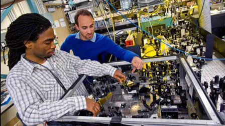 Photo of a black male scientist (left) and a white male scientist (right) working on a piece of electronic equipment in a National Laboratory. 